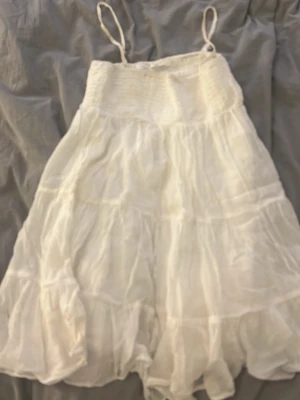 Vit sommarklänning med volanger - Luftig vit klänning med tunna axelband och volangdetaljer. Klänningen har smock upptill och är gjord i ett lätt, skirt tyg som passar perfekt för varma dagar.