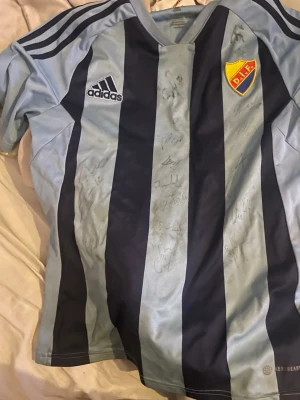 Blårandig Djurgården matchtröja Adidas - Säljer en blårandig matchtröja från Djurgården med Adidas-logga och klubbmärke på bröstet. Tröjan har flera autografer på framsidan och är tillverkad i ett lätt och ventilerande material, perfekt för fotboll eller samling.alla autografer från 2021