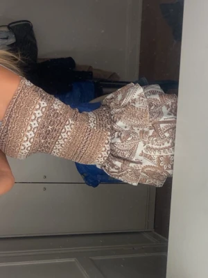 Brunmönstrad bandeauklänning - Kort bandeauklänning med brunt och vitt paisleymönster och smockad överdel. Kjolen har volanger och ger en bohemisk känsla. Perfekt för sommardagar och festivaler. Materialet ser ut att vara lätt och luftigt, troligen bomull eller viskos.