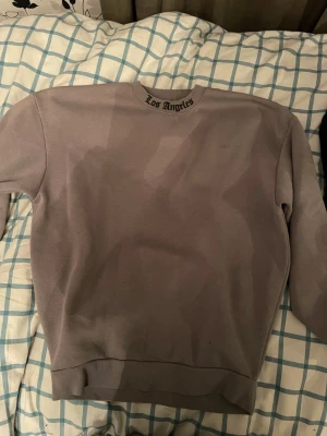 Ljusgrå sweatshirt Los Angeles - Ljusgrå oversized sweatshirt från Black Squad i storlek XS. Tröjan har rund halsringning, ribbade muddar och texten 'Los Angeles' broderad i nacken. Mjuk insida för extra komfort, perfekt för en avslappnad stil. Väldigt stor tröja för att vara en XS.