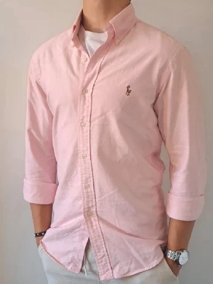 Rosa skjorta från Ralph Lauren - Klassisk rosa skjorta från Ralph Lauren med button-down krage och broderad logga på bröstet. Skjortan har långa ärmar, normal passform och stängs med vita knappar framtill. Perfekt för en clean och stilren look.