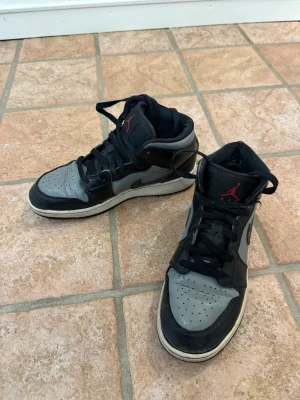 Air Jordan 1 Mid svart/grå - Säljer ett par Air Jordan 1 Mid i svart och grått läder med röda detaljer på plösen och sidan. Klassisk Nike Swoosh på sidan och perforerad tåbox för extra ventilation. Skorna har en hög siluett och svart snörning. Perfekta för dig som gillar streetwear.