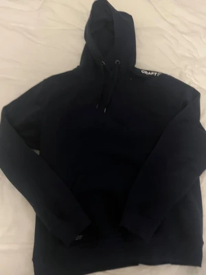 Mörkblå hoodie från Craft - Snygg mörkblå hoodie från Craft med dragsko i huvan och logga tryckt i vitt på axeln. Klassisk passform med långa ärmar och mjukt material som känns skönt mot huden. Perfekt för chill dagar eller när du vill ha en enkel och stilren look.