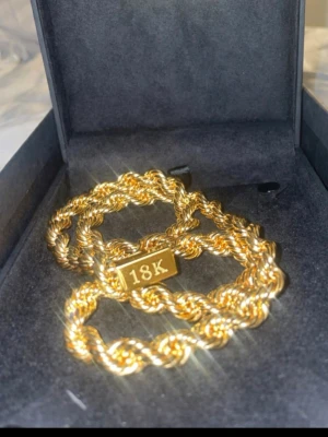 Guldhalsband 18K med tvinnad kedja - Snyggt halsband i guldfärg med tvinnad kedja och en bricka märkt 18K. Halsbandet har ett glittrande lås med små stenar för extra bling. Perfekt accessoar för att lyfta din stil.