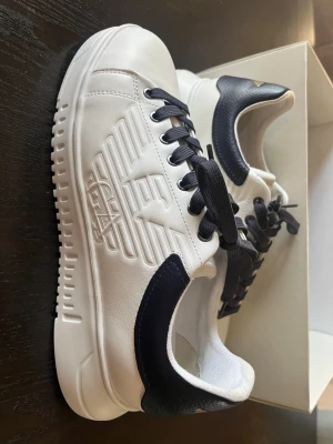 Vita sneakers från Emporio Armani - Kom med bud! Skriv gärna om ni har frågor eller vill diskutera pris. Stilrena vita sneakers från Emporio Armani med mörkblå hälparti och mörkblåa snören. Skorna har en präglad logga på sidan och är tillverkade i slätt läder. Snygg kontrast mellan vitt och mörkblått gör dem riktigt trendiga.