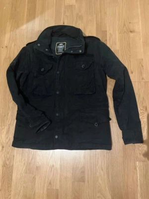Superdry field jacka - Svart parkajacka från Mountain Works med fyra stora fickor framtill och knappar längs hela framsidan. Jackan har hög krage och långärmad design, perfekt för kyligare dagar. Tillverkad i slitstarkt bomullsmaterial med klassisk passform.