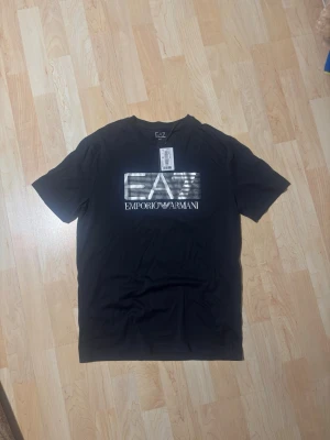 Svart EA7 Emporio Armani t-shirt - Snygg svart t-shirt från EA7 Emporio Armani med stort vitt logotryck på bröstet. Klassisk rund hals och korta ärmar. Perfekt för dig som vill ha en stilren och trendig look med exklusiv känsla.