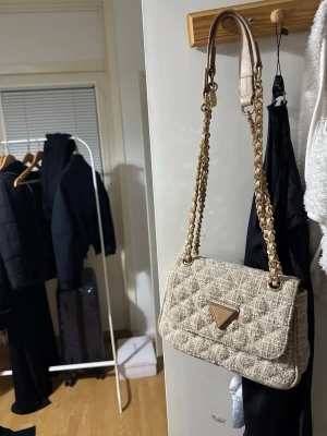 Beige quiltad handväska från Guess - Stilren beige handväska från Guess med quiltad struktur och guldiga kedjedetaljer på axelremmen. Väskan har en klassisk rektangulär form och är tillverkad i ett tygmaterial med tydlig textur. Guess-loggan i guld sitter framtill på väskan.