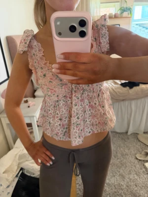 Blommig rosa croppad blus med rosett - Supersöt blommig blus i ljusrosa med volangdetaljer och knytband på axlarna. Croppad modell med v-ringning och spetsdetaljer, perfekt för sommaren. Tunn och luftig, passar till både jeans och kjol.