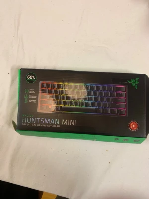 Razer Hunts evman Mini RGB tangentbord - Säljer ett Razer Huntsman Mini, ett kompakt 60% optiskt gamingtangentbord med RGB-belysning. Tangentbordet är svart med regnbågsfärgad bakgrundsbelysning och har en stilren, rektangulär form. Levereras i originalkartong med avtagbar USB-C kabel.