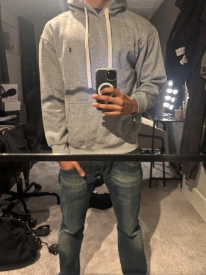 Grå Ralph Lauren Hoodie - Snygg grå hoodie från Polo Ralph Lauren med klassisk huva och vita dragsnören. Har en liten mörkblå logga broderad på bröstet och en stor magficka framtill. Tillverkad i mjukt bomullsmaterial som är perfekt för chill dagar. Storlek m men passar som s