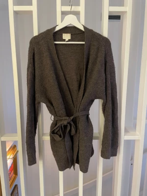 Brun ribbstickad kofta från H&M - Mysig brun ribbstickad kofta från H&M med knytskärp i midjan och långa ärmar. Perfekt att slänga på sig över en t-shirt eller topp för en avslappnad och stilren look.