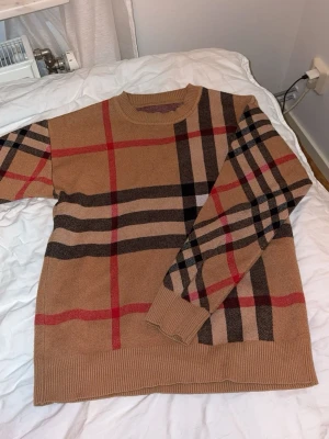 Burberry tröja  - Snygg beige stickad tröja med sopiga rutiga ränder i svart, rött och brunt. Tröjan har rund halsringning, långa ärmar och ribbade muddar vid ärmslut och nederkant. Perfekt för dig som gillar klassiska mönster och vill ha en trendig look.