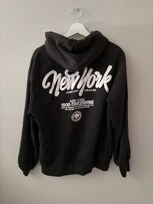 Svart hoodie med tryck  - Svart hoodie med stor vit New York-text och Time2Shine-tryck på ryggen samt mindre logga på bröstet. Klassisk känguruficka framtill och justerbar huva med snören. Mjuk och skön bomullsblandning, perfekt för en avslappnad streetwear-look.
