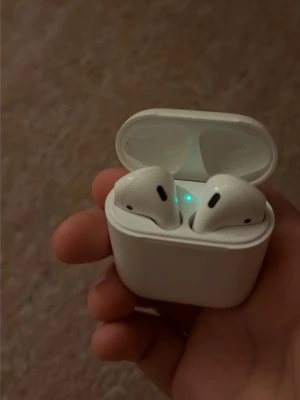 Apple AirPods med laddningsetui - Trådlösa vita in-ear hörlurar från Apple med klassisk stilren design. Kommer med ett kompakt laddningsetui som har LED-indikator på framsidan. Perfekta för musik, samtal och smidig anslutning till Apple-enheter.