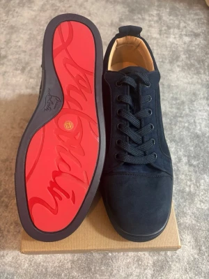 Marinblå sneakers från Christian Louboutin - Snygga marinblå sneakers i mocka från Christian Louboutin med klassisk röd sula och svarta skosnören. Låg modell med stilren design och exklusiv känsla. Perfekta för dig som vill sticka ut med en ikonisk detalj under skon.