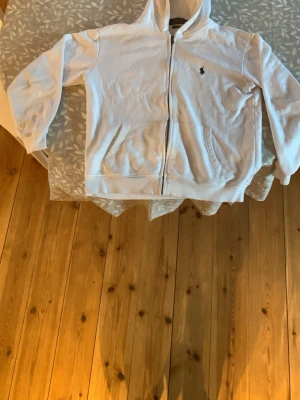 Vit hoodie sopolo Ralph sopolo - Säljer en vit hoodie från Ralph Lauren med dragkedja och sopolo-logga broderad på bröstet. Hoodien har huva och två fickor framtill. Tillverkad i mjukt material som känns skönt mot huden. Perfekt för en chill och avslappnad stil.