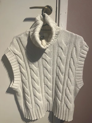 Vit kabelstickad väst från H&M - Säljer en krämvit kabelstickad väst från H&M med hög polokrage och breda ribbade kanter. Västen är ärmlös och har ett chunky stickat mönster som ger en trendig look. Perfekt att styla över en skjorta eller långärmad tröja.