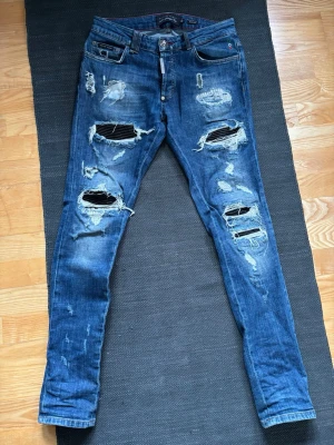 Blå slitna jeans från Philipp Plein - Säljer ett par blå jeans från Philipp Plein med kraftiga slitningar och hål på både fram- och baksida. Skinny fit-modell med klassiska fem fickor och detaljer som trasiga partier och råa kanter. Perfekta för en edgy streetstyle-look.