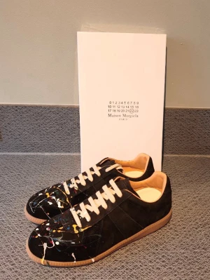 Maison Margiela svart graffiti  - Maison Margiela sneakers i svart mocka och läder med unika färgstänk i vitt, gult och rött. Klassisk låg modell med vita snören och beige innersula. Snygg kontrast mellan material och färgdetaljer, perfekt för dig som gillar statement-plagg.