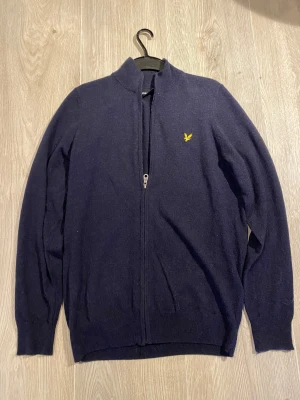 Mörkblå ziptröja från Lyle & Scott - Mörkblå ziptröja från Lyle & Scott med gul broderad logga på bröstet. Tröjan har hög krage, hel dragkedja och ribbade muddar vid ärmslut och nederkant. Perfekt för lager-på-lager och enkel att matcha. Tröjan är helt ny och inte använd.