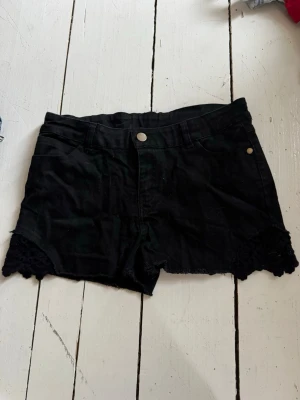 Svarta shorts med spetsdetalj - Snygga svarta shorts med spetsdetalj längst ner på benen. Klassisk femficksmodell med knapp och dragkedja framtill. Perfekta för varma dagar och ger en trendig look.
