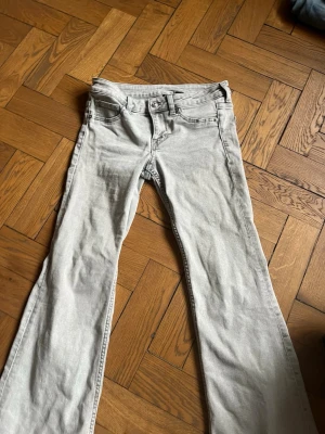 Ljusgrå bootcut jeans - Säljer ett par ljusgrå low waist bootcut jeans med klassisk femficksdesign och låg midja. Jeansen har utsvängda ben och är tillverkade i denim med en mjuk känsla. Perfekta för dig som gillar en avslappnad och trendig look.