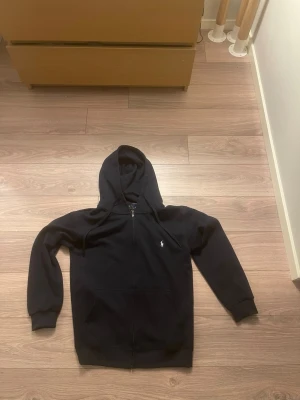 Svart hoodie från Polo Ralph Lauren - Pris går att diskutera på en snygg svart hoodie från Polo Ralph Lauren med dragkedja framtill och klassisk vit logga broderad på bröstet. Hoodien har huva med dragsko och två fickor framtill. Tillverkad i mjukt bomullsmaterial som känns skönt mot huden.