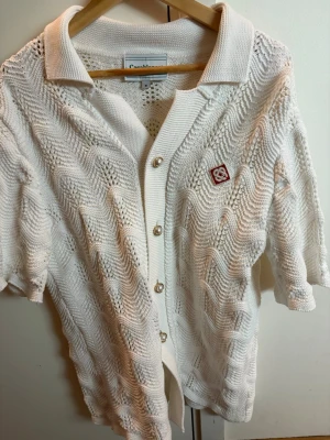 Vit stickad skjorta från Casablanca  - Vit stickad skjorta från Carhartt med korta ärmar och klassisk krage. Skjortan har ett snyggt hålmönster, pärlemorknappar framtill och en liten broderad logga på bröstet. Perfekt för en avslappnad och trendig stil.