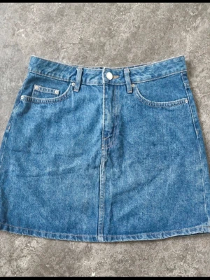 SOM NY Blå jeanskjol med fickor från DIVIDED stl 36 - Klassisk blå jeanskjol i kort modell med fem fickor och bälteshällor. Kjolen har knapp och dragkedja framtill och är gjord i ett tåligt denimtyg. Perfekt basplagg för en avslappnad och trendig stil. Jeanskjol stl 36 Från Divided Längd ca 40 cm Midjemått 35 cm ( 70 cm runt ) Använd 1 g URSÄKTA EN SKRYNKLIG BILD