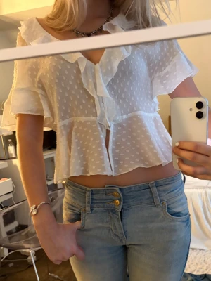 Vit volangblus med prickigt mönster - Söt vit blus med volangkant och prickigt mönster. Blusen har korta ärmar, djup v-ringning och knappar framtill. Tillverkad i ett lätt, transparent tyg som ger en luftig känsla. Man kan både ha den stäng och knäppa upp den med knapparna