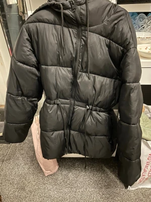 Svart pufferjacka med huva - Säljer en svart pufferjacka med huva och dragkedja framtill. Jackan har justerbar midja med snörning och stora puffiga sektioner som ger en riktigt trendig look. Perfekt för kalla dagar och riktigt varm. Materialet är glansigt och vattenavvisande.