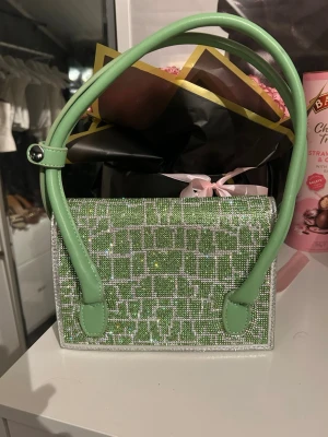 Grön glittrig handväska var med pärlor - Trendig grön handväska med glittrande pärlor och rutmönster. Väskan har dubbla gröna handtag i konstläder och en rektangulär form. Perfekt accessoar för att lyfta din outfit med lite extra bling.