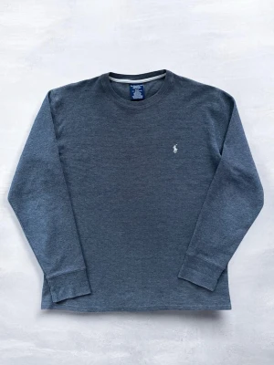 Mörkgrå tröja från Polo Ralph Lauren - Säljer en mörkgrå tröja från Polo Ralph Lauren med rund halsringning och diskret vit logobrodyr på bröstet. Tröjan är stickad i ett mjukt material och har en klassisk passform som är enkel att matcha.