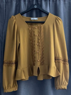 Senapsgul blus med spetsdetaljer - Trendig senapsgul blus från Hare med långa puffärmar och dekorativa spetsdetaljer längs ärmar och framtill. Blusen har klädda knappar och volang nedtill, samt fyrkantig ringning för en cool vintagekänsla. Perfekt för dig som gillar unika plagg.