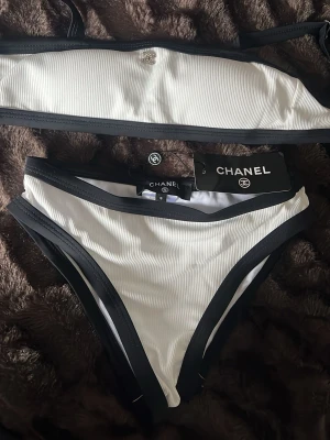 Vit och svart bikini från Chanel - Snygg bikini från Chanel i vit ribbad design med svarta kanter. Både överdel och underdel har diskret Chanel-logga och stilrena detaljer. Perfekt för sommarens strandhäng och poolpartyn. Materialet är syntet och ger en bekväm passform. Helt ny 