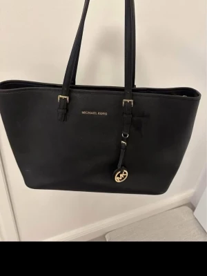 Svart Michael Kors handväska - Stilren svart handväska från Michael Kors i slätt läder med guldfärgade detaljer och dubbla axelband. Väskan har ett klassiskt MK-hänge och diskret logga framtill. Perfekt för dig som gillar en enkel men lyxig look. Köpt på zalando för några år sen, fint skick