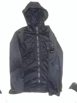 Svart vindjacka från C.P. Company - Snygg svart vindjacka från C.P. Company med huva och dragkedja framtill. Jackan har två bröstfickor med knappar och den ikoniska linsen på ena fickan. Tillverkad i ett lätt, vindtätt material som passar perfekt till streetwear.