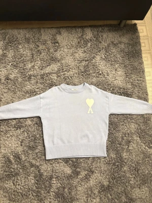 Ljusgrå stickad tröja från Ami Paris - Supersnygg ljusgrå stickad tröja från Ami Paris med ikoniskt hjärta och A på bröstet. Tröjan har rund hals, ribbade muddar och en croppad passform som ger en modern vibe. Perfekt till jeans för en clean look.