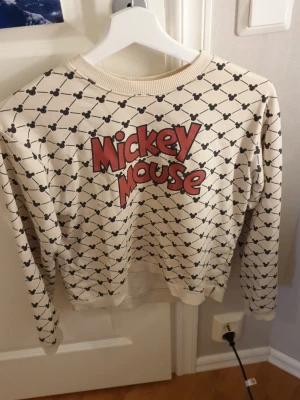 Mickey Mouse sweatshirt från H&M - Cool beige sweatshirt från H&M med svart Mickey Mouse-mönster över hela tröjan och stor röd text framtill. Rund halsringning och ribbade muddar. Perfekt för dig som gillar Disney och vill ha en unik och bekväm tröja.