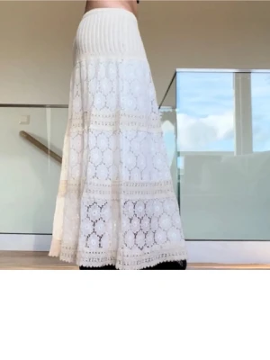 Vit långkjol i spets med volang - Superfin vit långkjol med spetsdetaljer och volang. Kjolen har ett ribbat midjeband och vackert broderat mönster över hela tyget. Perfekt för dig som gillar romantisk och bohemisk stil.