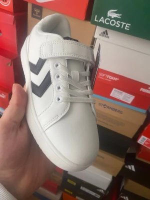 Vita Hummel sneakers med kardborre - Snygga vita sneakers från Hummel med svarta chevron-detaljer på sidan. Skorna har både snörning och kardborreband för enkel påtagning. Tillverkade i syntetmaterial med klassisk låg profil och vit sula. Perfekta för en clean och sportig look.