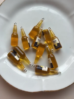 Miniatyr ölflaska smyckesberlocker - Säljer flera små smyckesberlocker formade som ölflaskor med gul färg och etikett som liknar Corona. Perfekta att använda till halsband, armband eller örhängen. Tillverkade i plast med metallögla för enkel montering. Rolig detalj för dig som gillar unika accessoarer.
