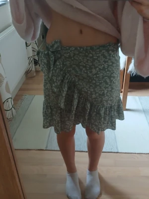 Grön blommig omlottkjol med volang - Supersöt grön omlottkjol med vitt blommönster och volangkant. Kjolen har en knytning i midjan som ger en snygg detalj och sitter skönt på kroppen. Perfekt för dig som gillar en feminin och trendig stil.