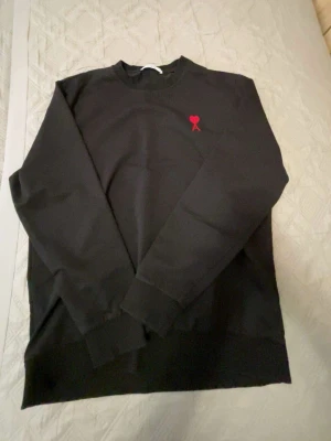 Svart sweatshirt med rött hjärta - Svart sweatshirt med rund hals och ett litet rött hjärta broderat på bröstet. Tröjan har långa ärmar och ribbade muddar vid ärmslut och nederkant. Perfekt för en enkel och stilren look.