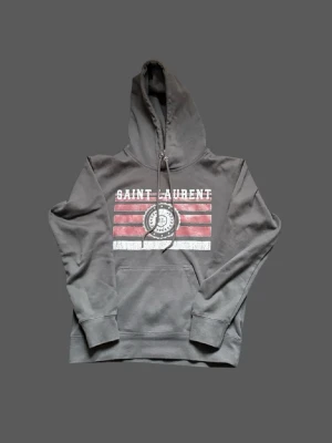 Grå Saint Laurent hoodie med tryck - Snygg grå hoodie från Saint Laurent med huva och dragsko. Framsidan har ett stort tryck med texten 'SAINT LAURENT' och röda samt vita ränder, plus ett runt emblem med 'YSL'. Klassisk känguruficka och ribbade muddar. Perfekt för en avslappnad streetwear-look. Den är lite liten i storlek sitter som xs.