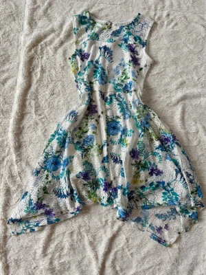 Vit blommig spetsklänning - Säljer en vit ärmlös klänning med blått, grönt och lila blommönster. Klänningen har spetsdetaljer och asymmetrisk nederkant som ger en unik look. Perfekt för sommarens alla tillfällen och har en luftig passform.
