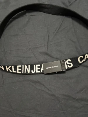 Svart bälte från Calvin Klein Jeans - Säljer ett svart bälte från Calvin Klein Jeans med vit text längs hela bandet. Bältet har en enkel metallspänne och är tillverkat i syntetmaterial. Perfekt för att ge din outfit en cool touch med tydlig logga.