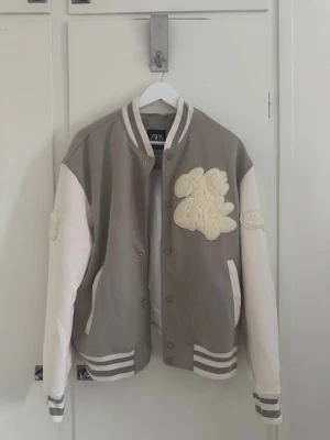 Baseballjacka från Zara - Snygg baseballjacka från Zara i grått och vitt med broderade detaljer på bröstet och ärmen. Jackan har ribbade muddar med ränder, tryckknappar framtill och två fickor. Oversized passform och klassisk college-look som ger en trendig vibe. Den är aldrig använd.