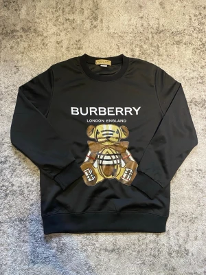 Svart Burberry sweatshirt med björn - Cool svart sweatshirt från Burberry med stort tryck av en nallebjörn i klassiskt rutmönster och texten 'BURBERRY LONDON ENGLAND' på bröstet. Tröjan har rund hals, långa ärmar och ribbade muddar. Storleken är XS-S och passar dig runt 165-170 lång. Hör gärna av dig vid frågor!